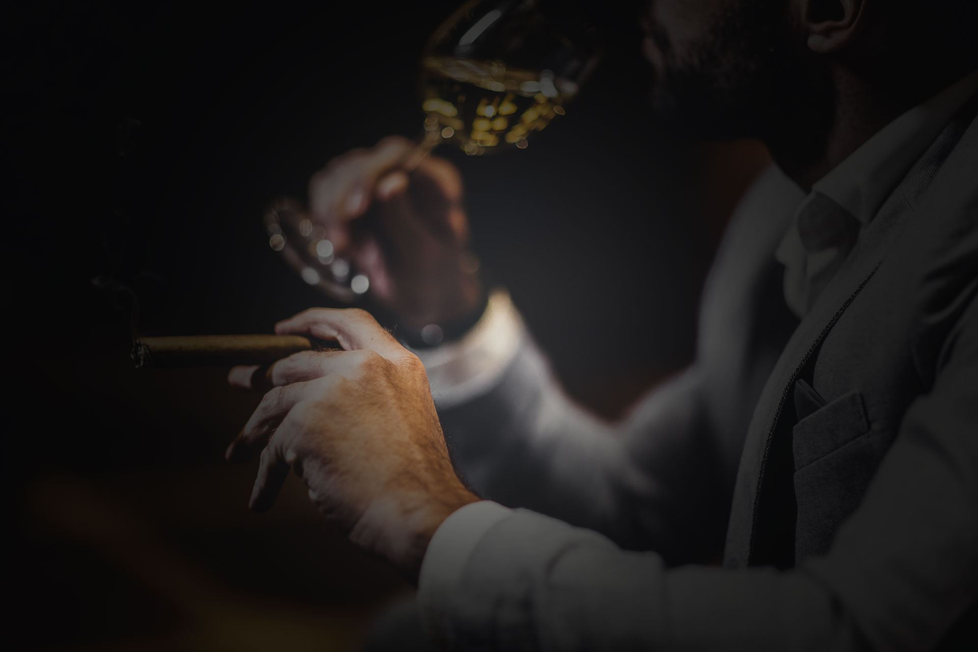 A man in a grey suit holding a cigar and a glass of whisky in low light